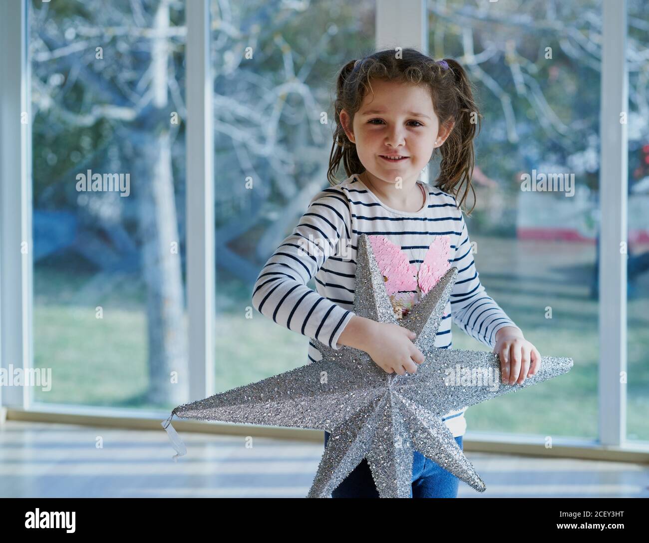 Smiling little girl with ponytails dressed in casual clothes holding silver star shaped Christmas decoration while standing against window at home Stock Photo