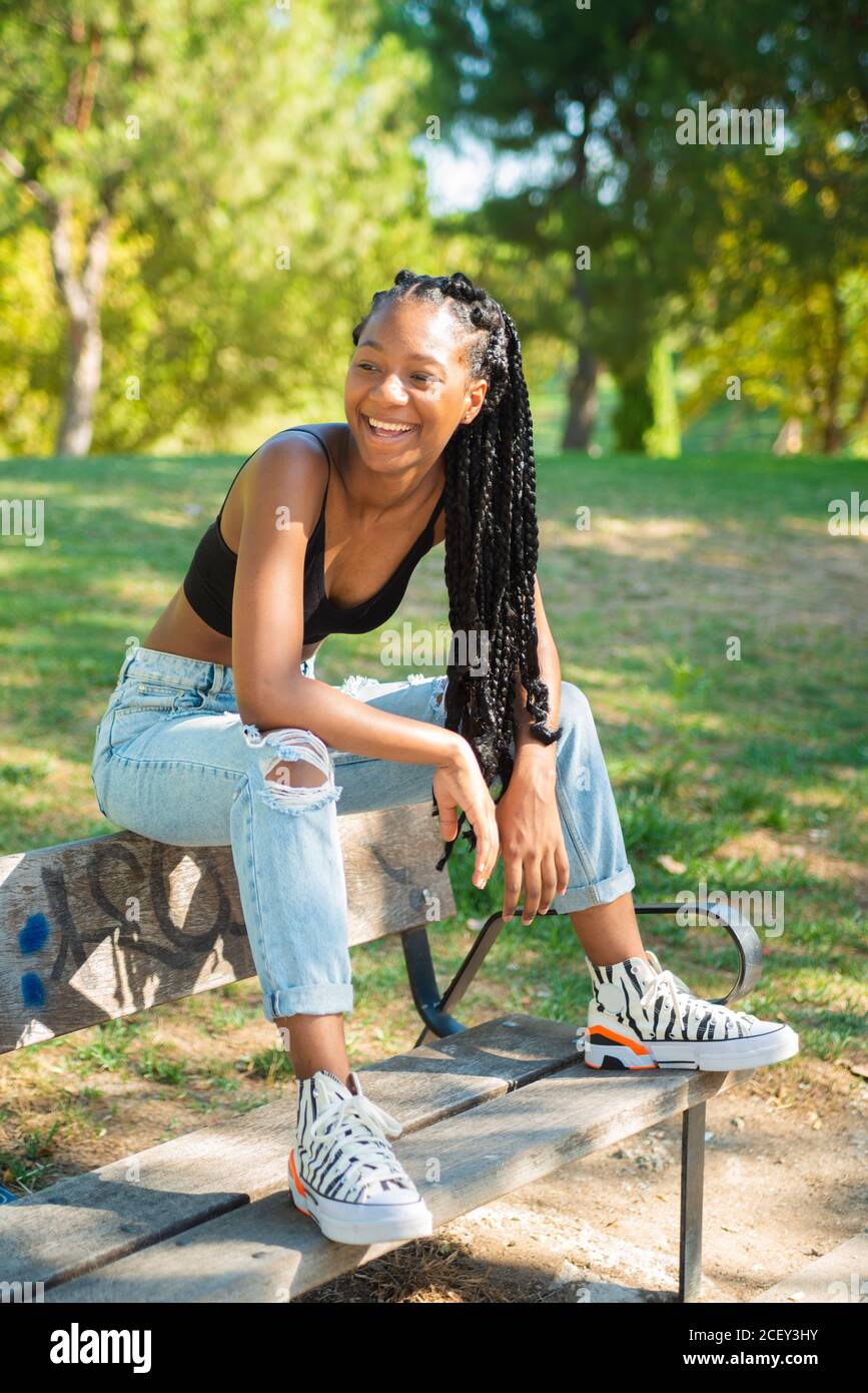 Full body of relaxed young African American female hipster in trendy outfit  sitting on wooden bench in green park in summer day Stock Photo - Alamy