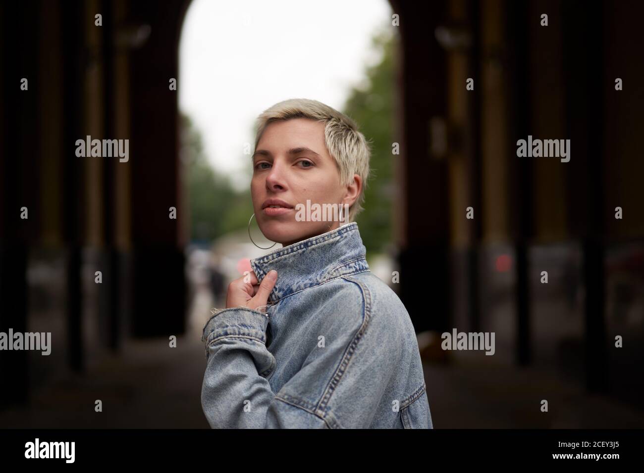 Side view of calm female model wearing denim jacket standing in arched passage on street and looking at camera Stock Photo