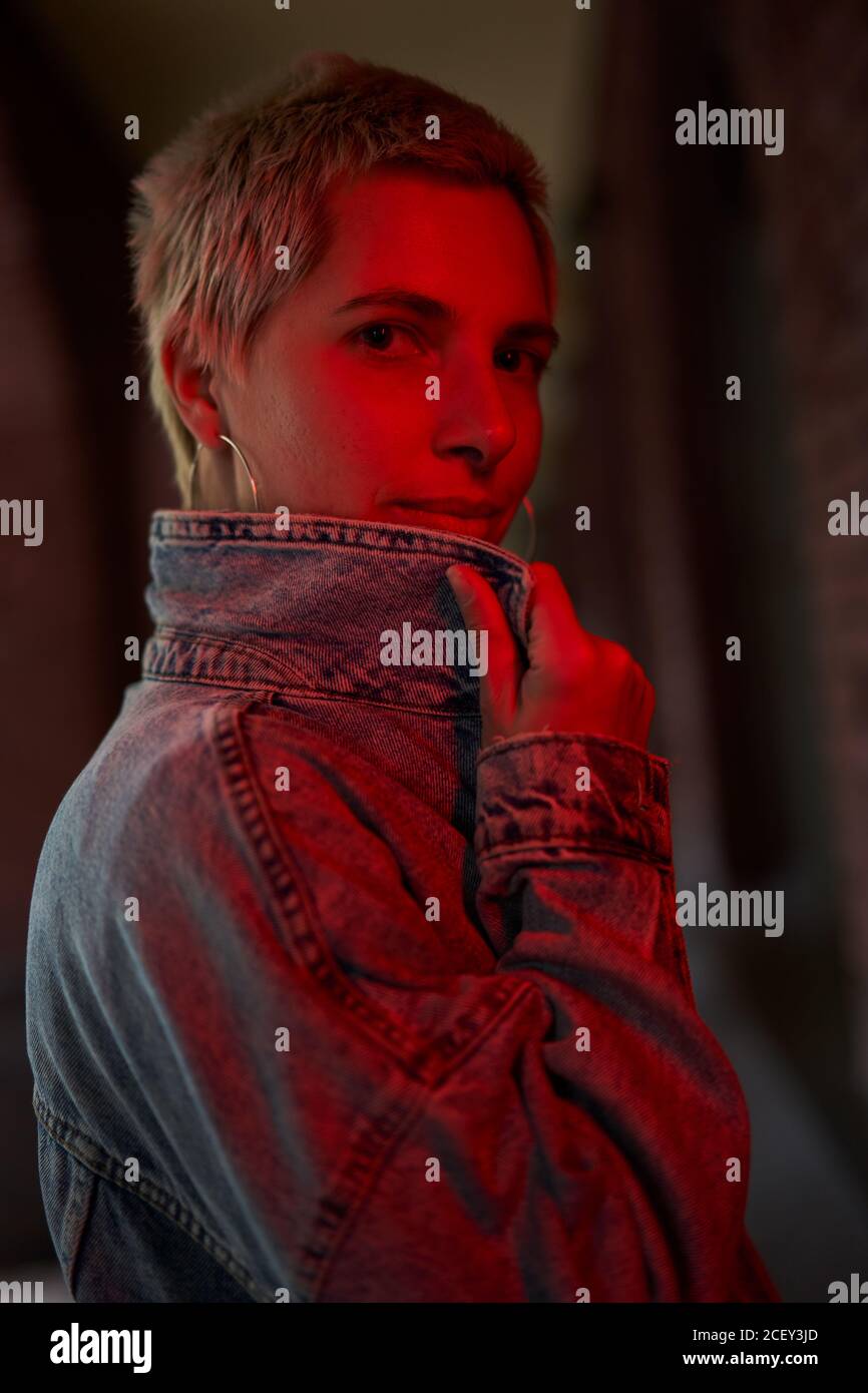 Side view of unemotional female with short hair and in denim jacket standing on street and illuminated by red light while looking at camera Stock Photo