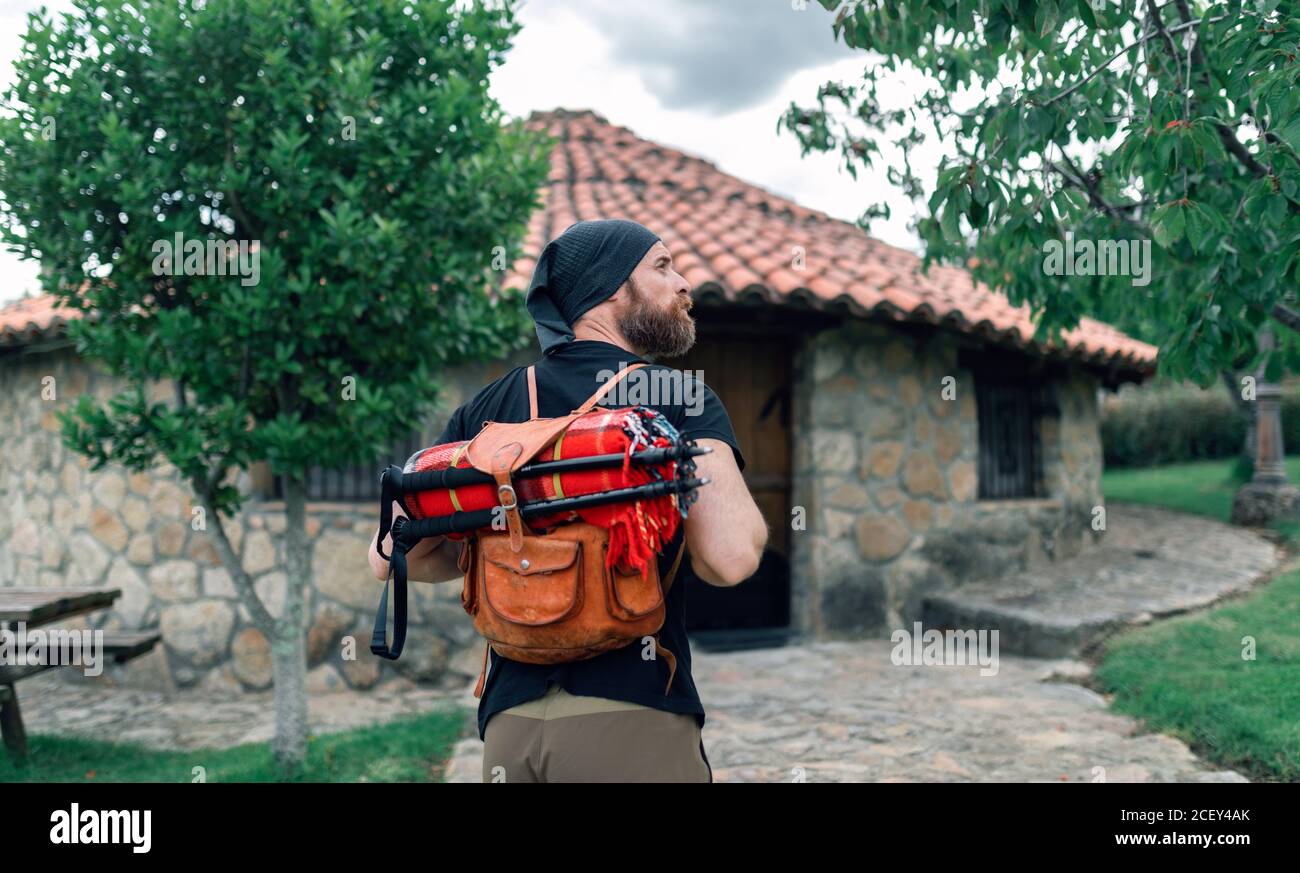 Back view of calm male tourist with backpack standing near stone house in settlement and looking away Stock Photo
