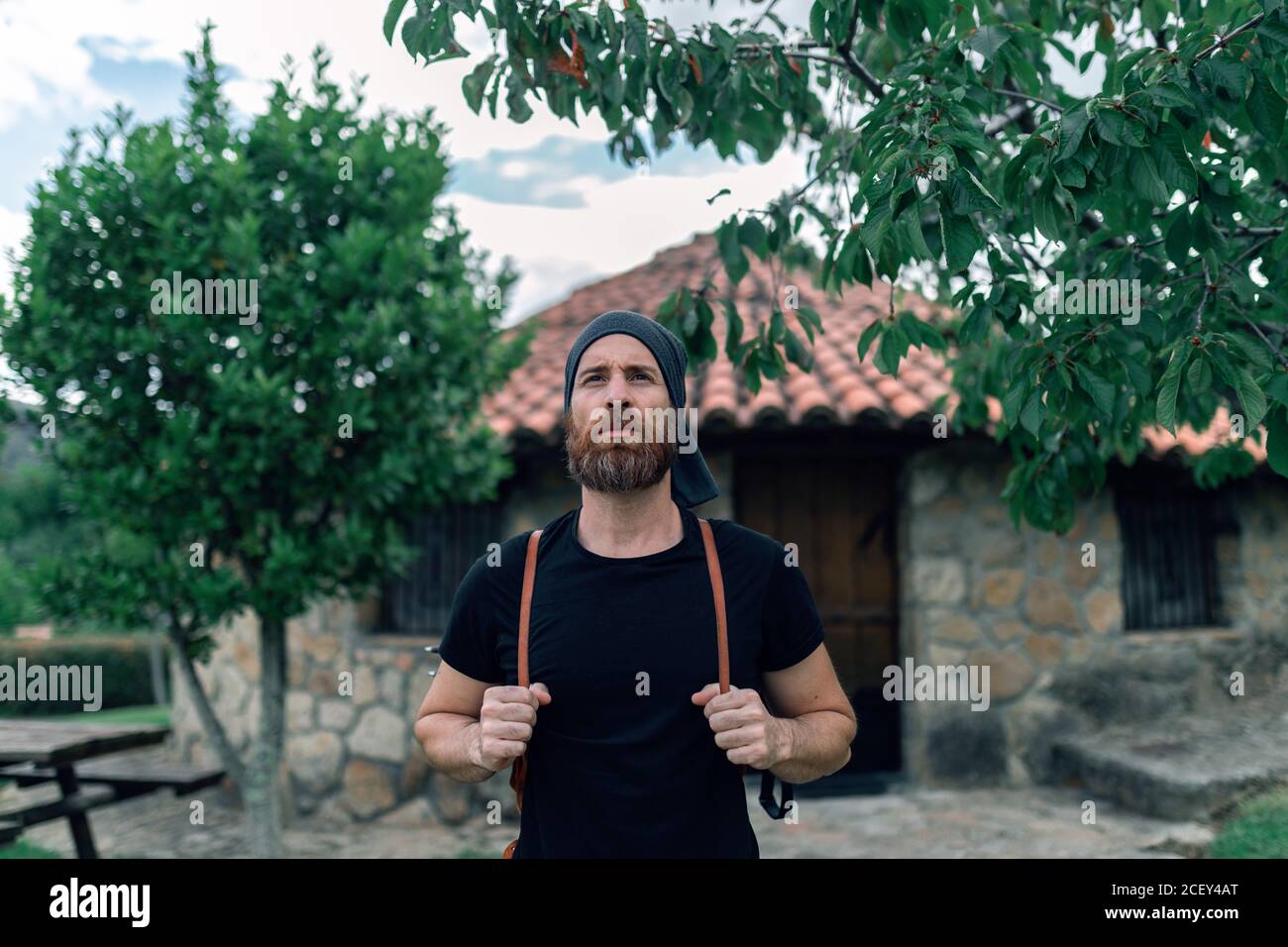 Calm male tourist with backpack standing near stone house in settlement and looking away Stock Photo