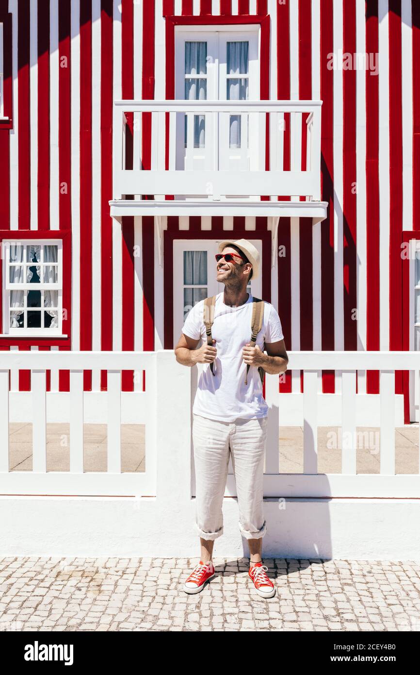 Full body happy guy in white outfit standing with backpack against colorful striped house on Costa Nova Stock Photo