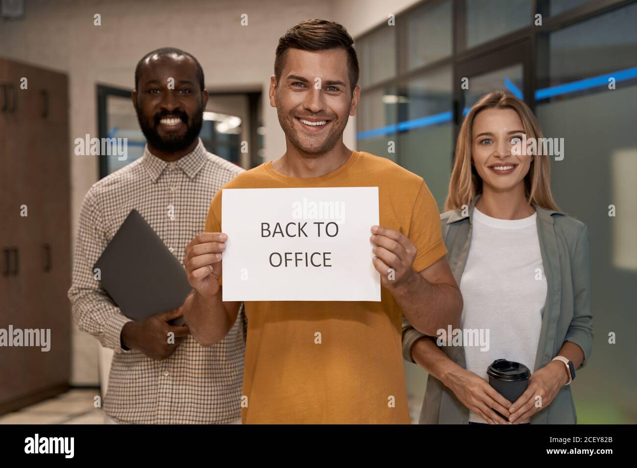 Young happy caucasian man, office worker showing paper with text BACK TO OFFICE at camera and smiling while standing with colleagues in the office Stock Photo