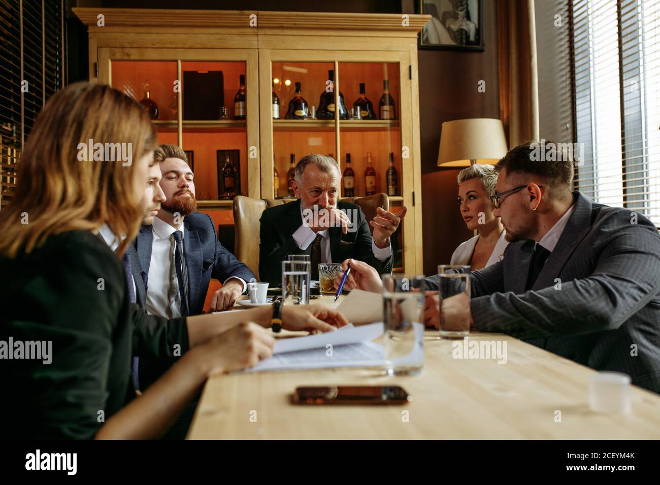 Aged boss sitting at head of wooden table in leather brown chair and  pointing to young team member in glasses. Business partners watching out  for lead Stock Photo - Alamy