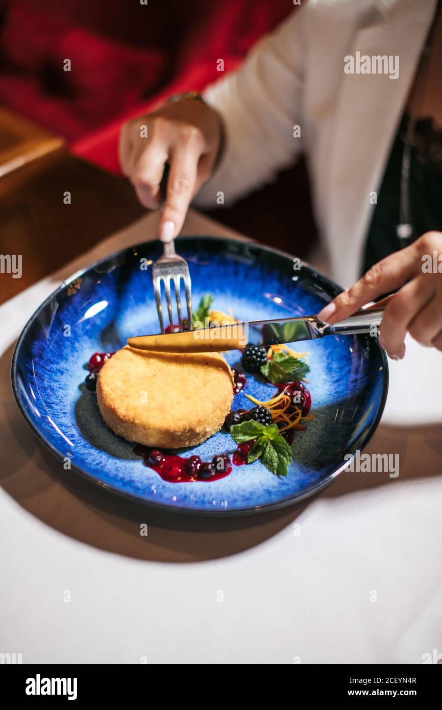 Delicate female hands holding silver fork and knife, cutting fresh prepared dessert, beautiful cake is served at table in glorious dark blue plate pre Stock Photo
