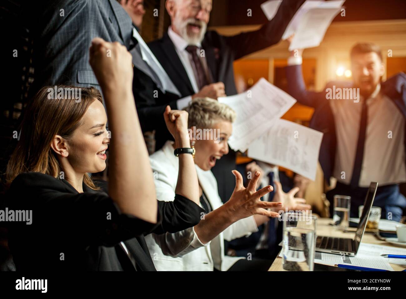Group of professionals enthusiastically celebrating good deal consummation, raising hands up, joyfully loudly yawling in business office Stock Photo