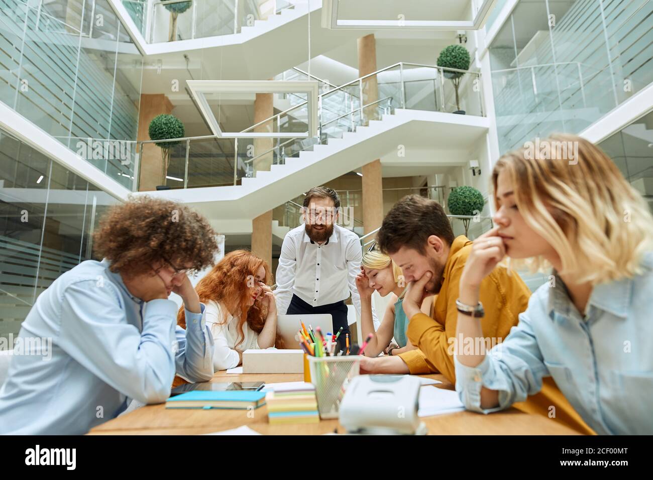 Elegant noble businessman with thick beard and round glasses, dressed in stylish white shirt leans on table, talks to smart employees with positive ex Stock Photo