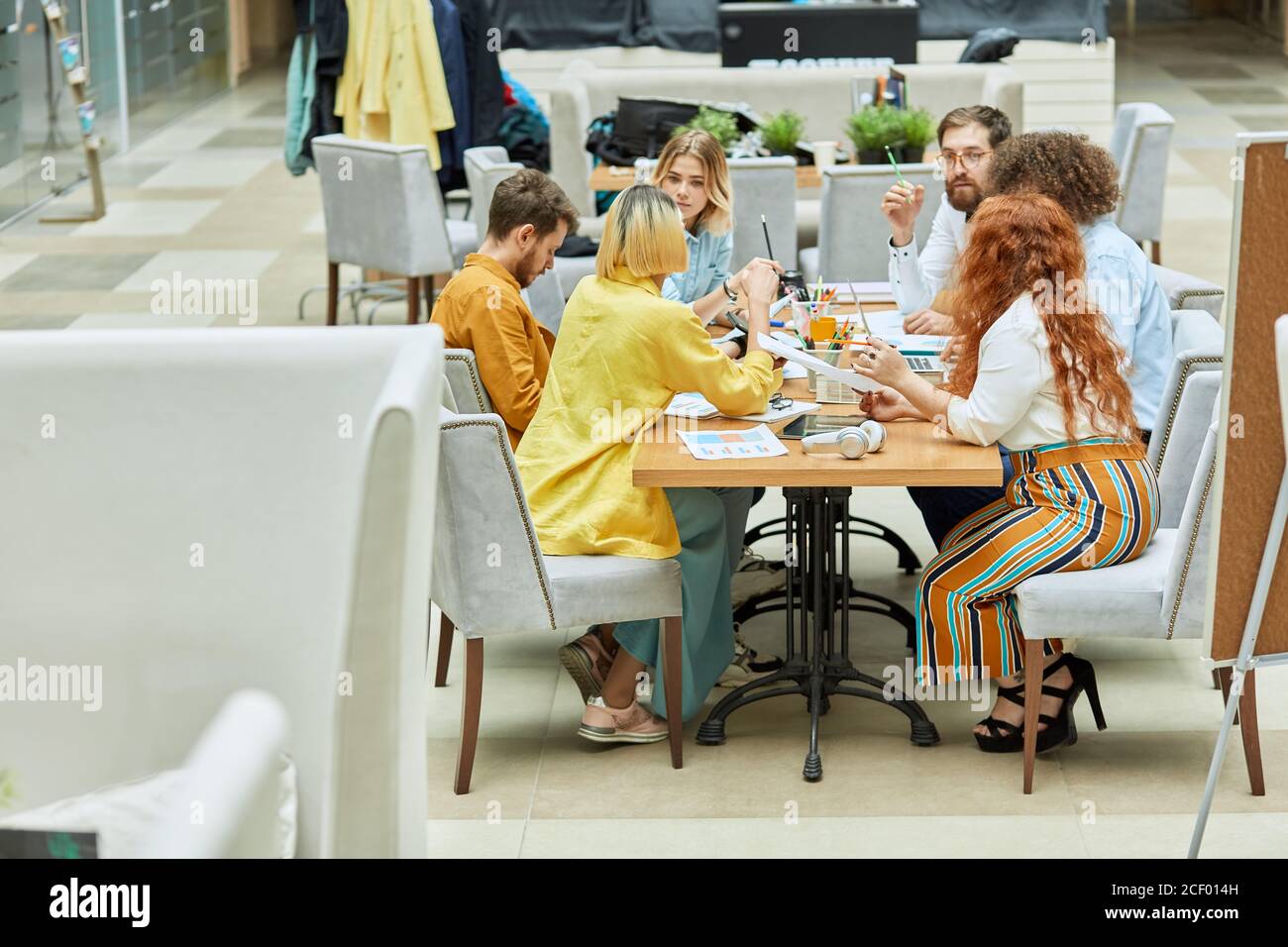 Group of concentrated busy graphic designers thinking over new project, siting at table full of paper and stationary items, brainstorming concept Stock Photo