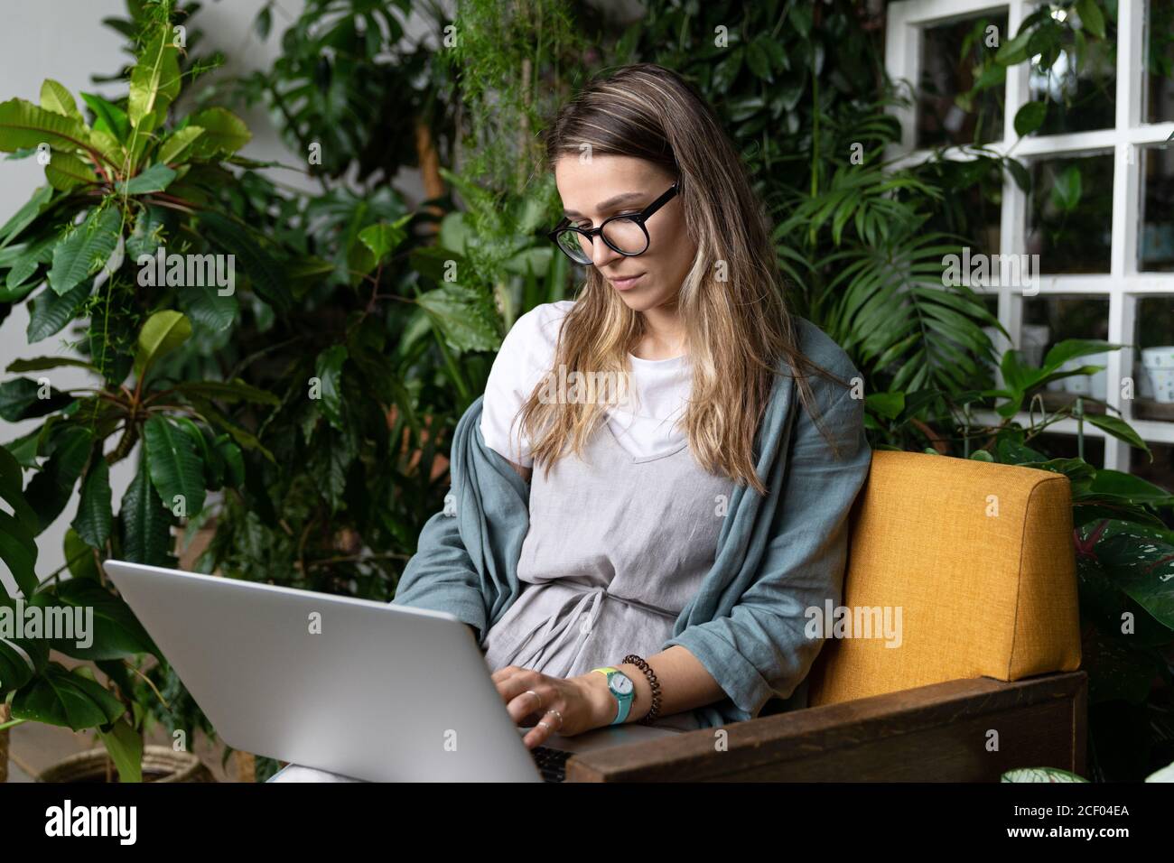 Woman gardener wear linen dress, sitting on chair in green house, working on laptop surrounded by plants. Home garden, remote work, distance job. Stock Photo