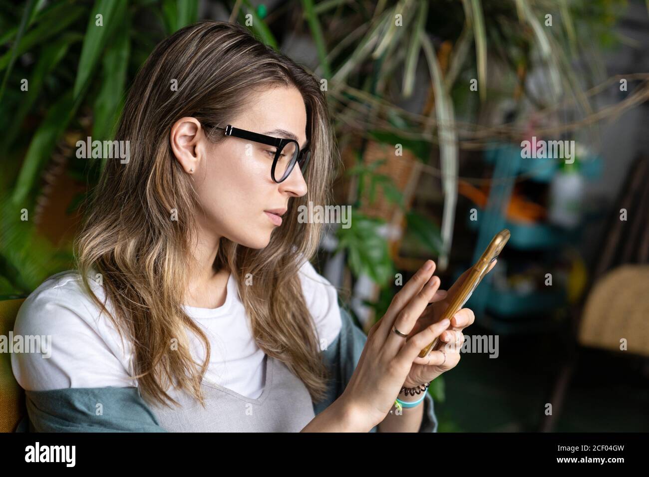 Close up of woman freelancer in green house, resting, using smartphone and sending message surrounded by exotic plants. Social network. Stock Photo