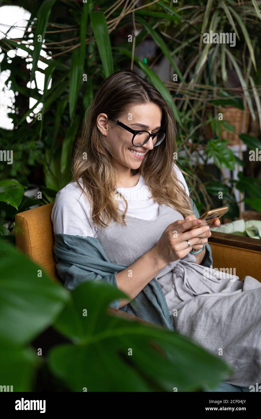 Close up of smiling positive female gardener wear linen dress, sitting on chair in green house, resting, using smartphone and sending message Stock Photo