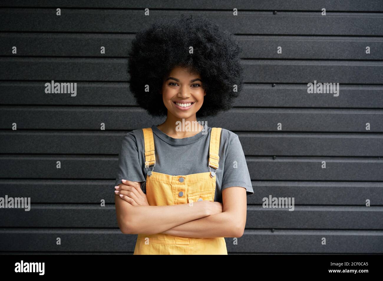 Smiling African hipster lady standing arms crossed against black wall. Stock Photo