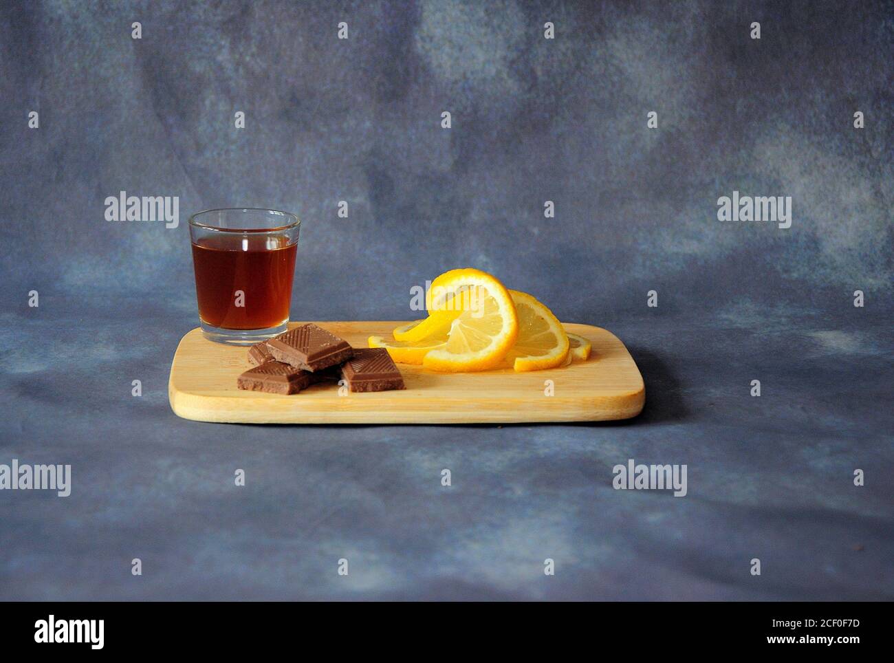 A glass of cognac, pieces of chocolate and lemon wedges lie on a wooden board on a gray background. Close-up. Stock Photo