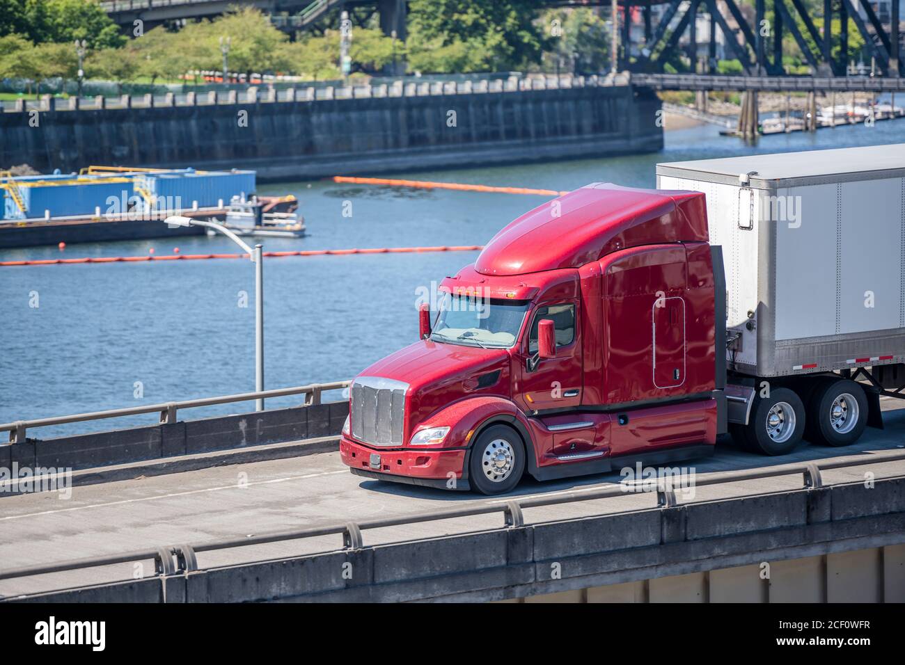 Red industrial grade diesel big rig semi truck with low cab for best aerodynamics transporting commercial cargo in dry van semi trailer running along Stock Photo