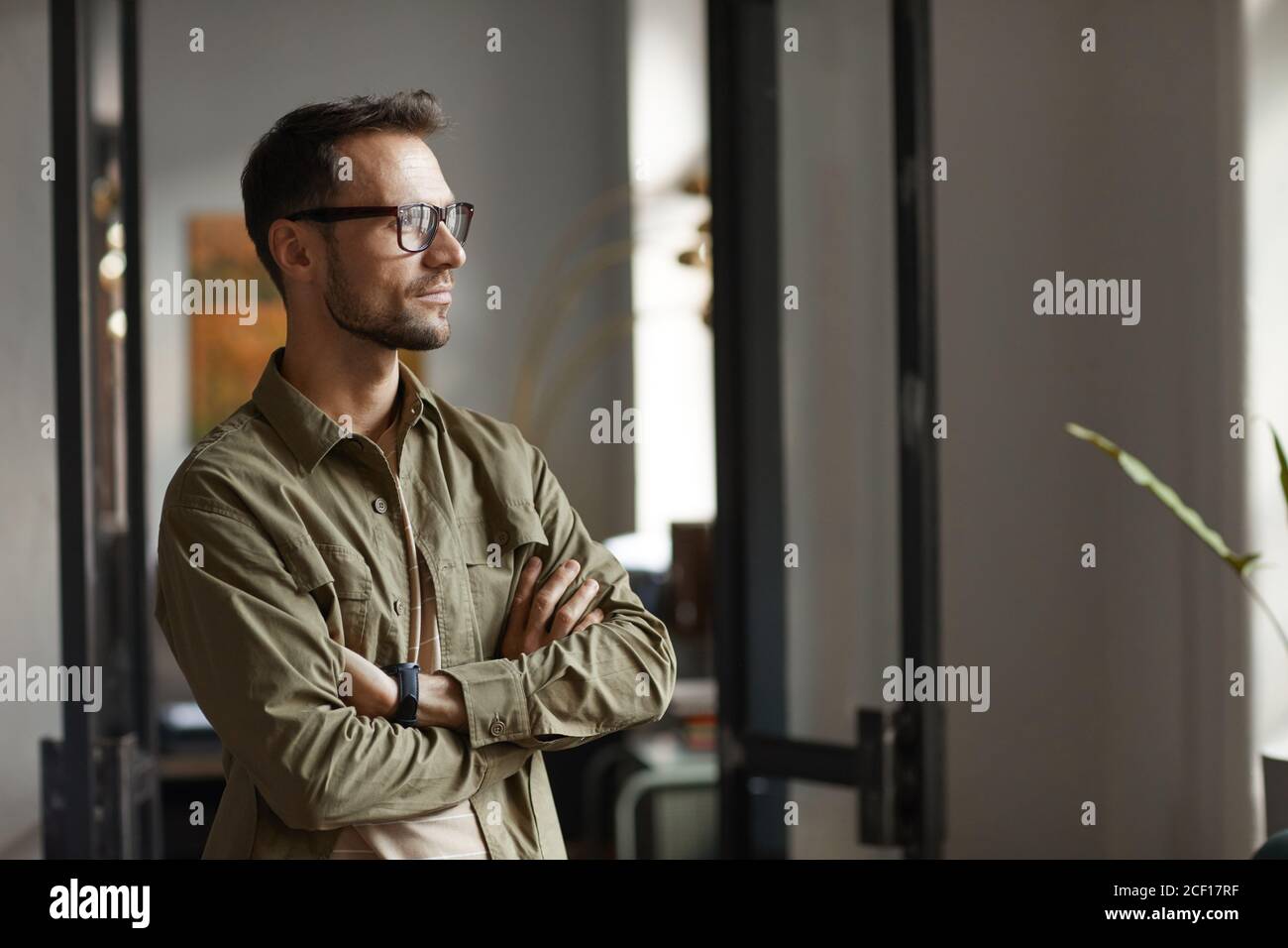 Young man in eyeglasses standing at office and looking through the window with pensive look Stock Photo