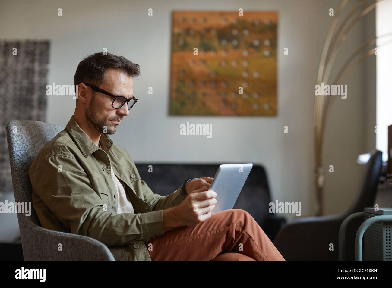 Serious designer in eyeglasses sitting on sofa and working online on digital tablet at the modern office Stock Photo