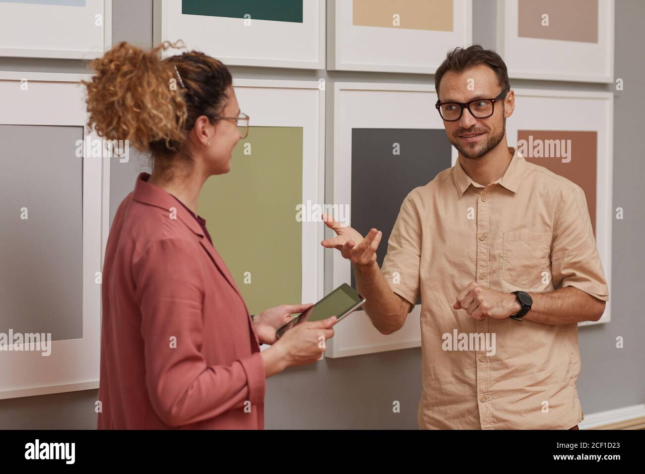 Young man talking to woman while she using tablet pc they standing against the pictures on the wall Stock Photo