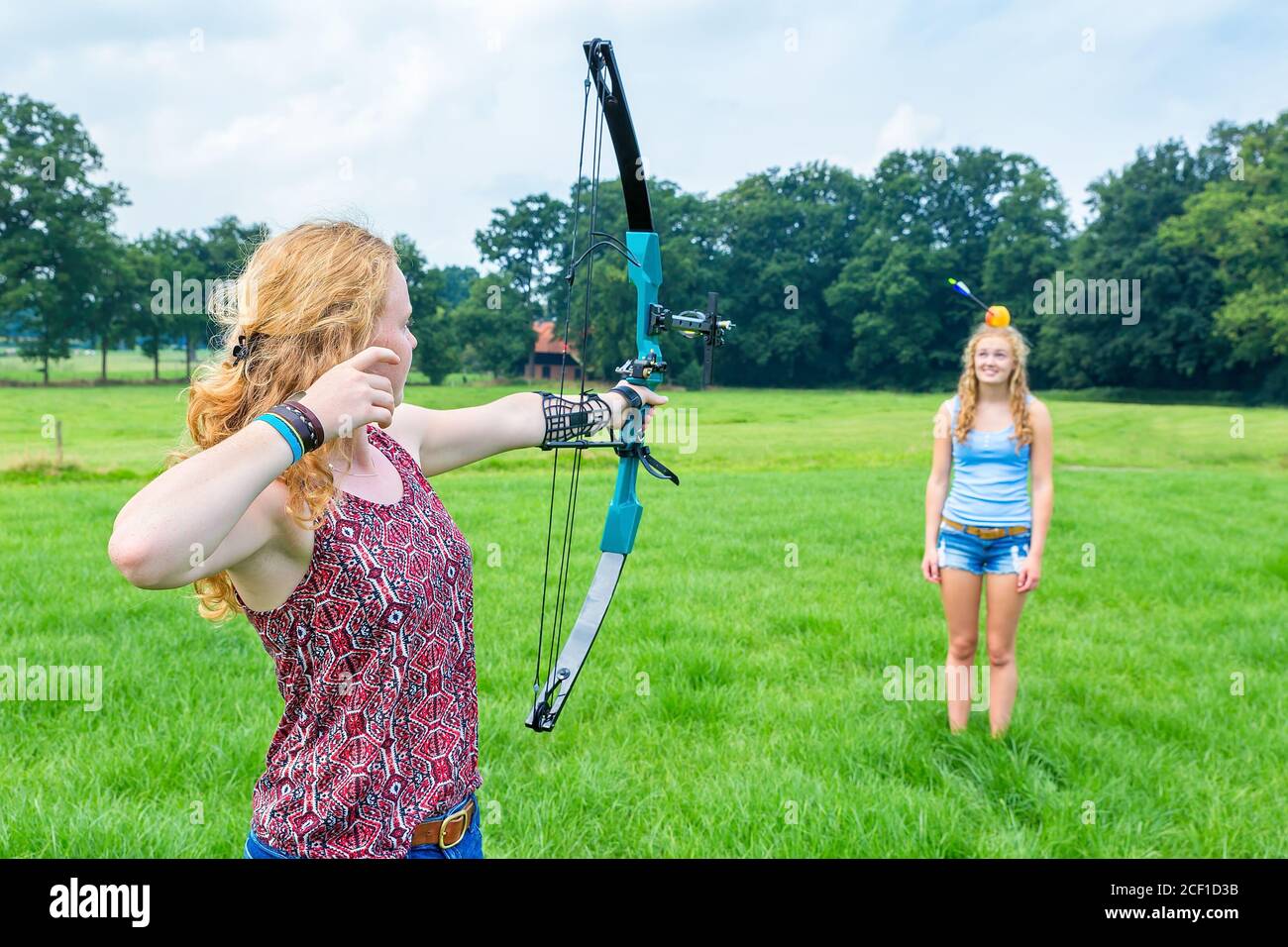 Dutch teenage girl aiming arrow of compound bow at fruit on head of young woman Stock Photo