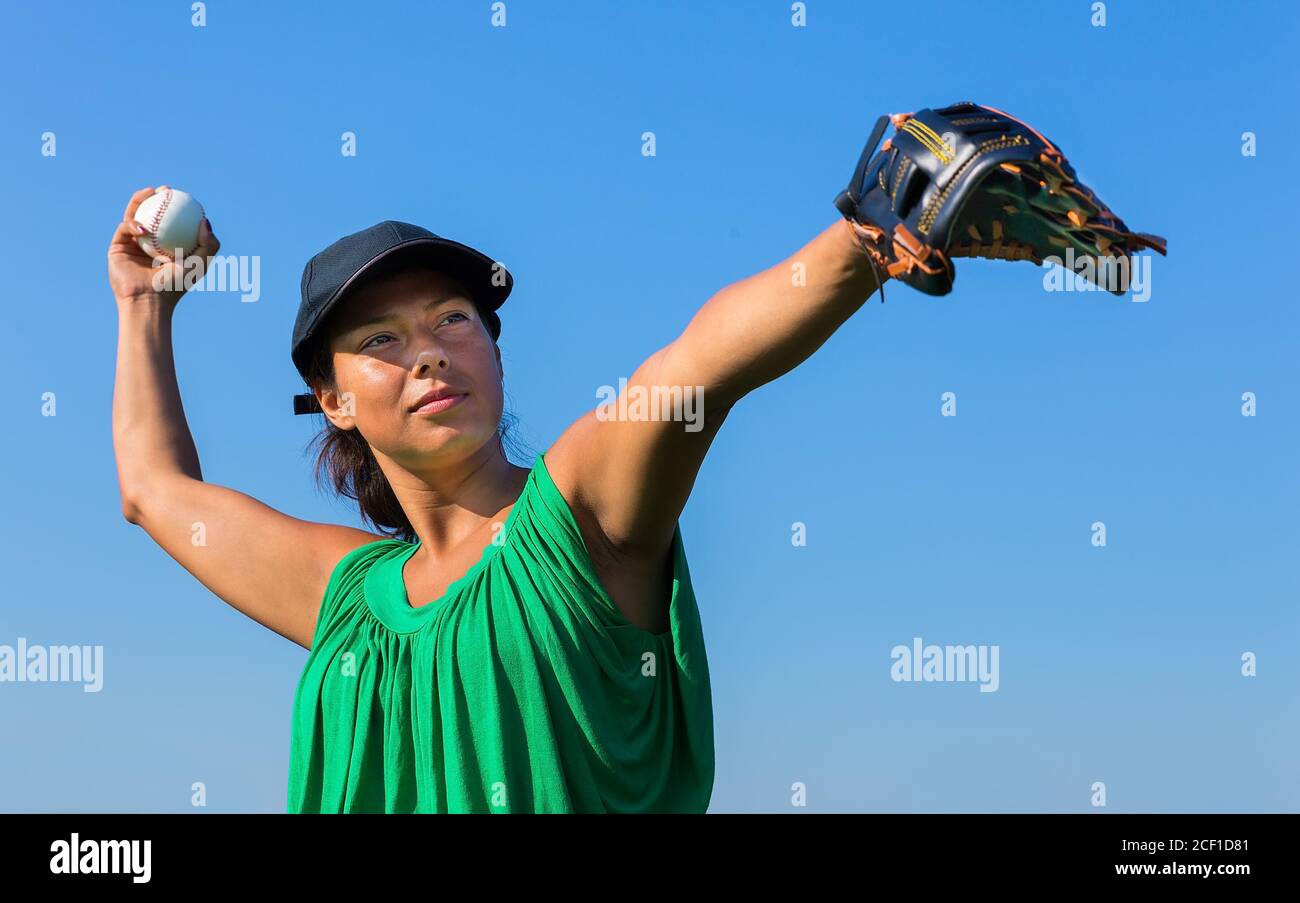 Colombian woman with baseball glove and cap throwing baseball in blue sky Stock Photo