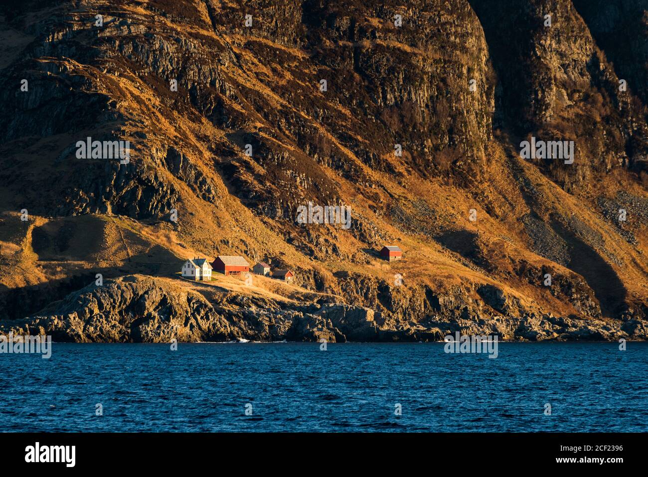 Steep cliff on the coast with small houses in barren landscape in Norway on bright sunny day seen from boat Stock Photo