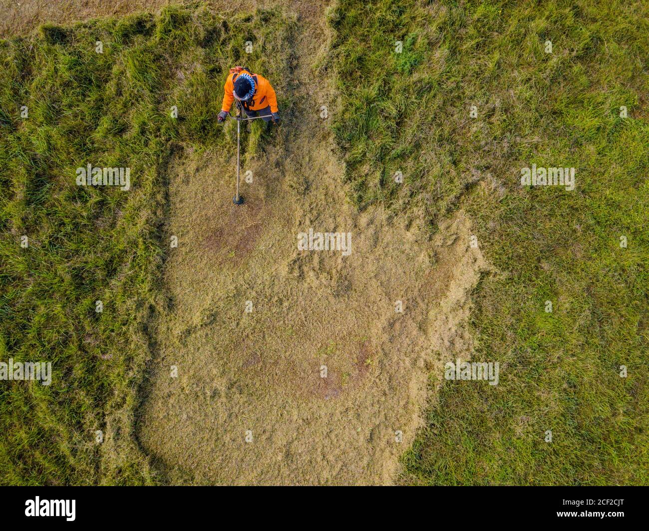 Aerial view of a man wearing protective clothing and eye covering, cutting a field of long grass. Stock Photo