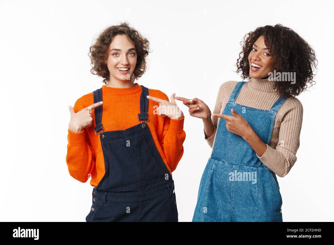 Portrait of two joyful women in overalls smiling and pointing fingers ...