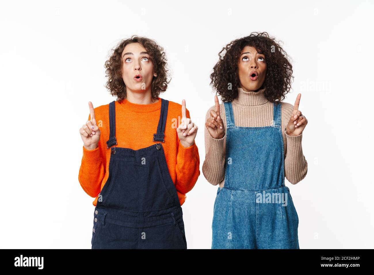 Image of shocked two multiracial girls friends in denim jumpsuits isolated over white wall background pointing to copyspace. Stock Photo