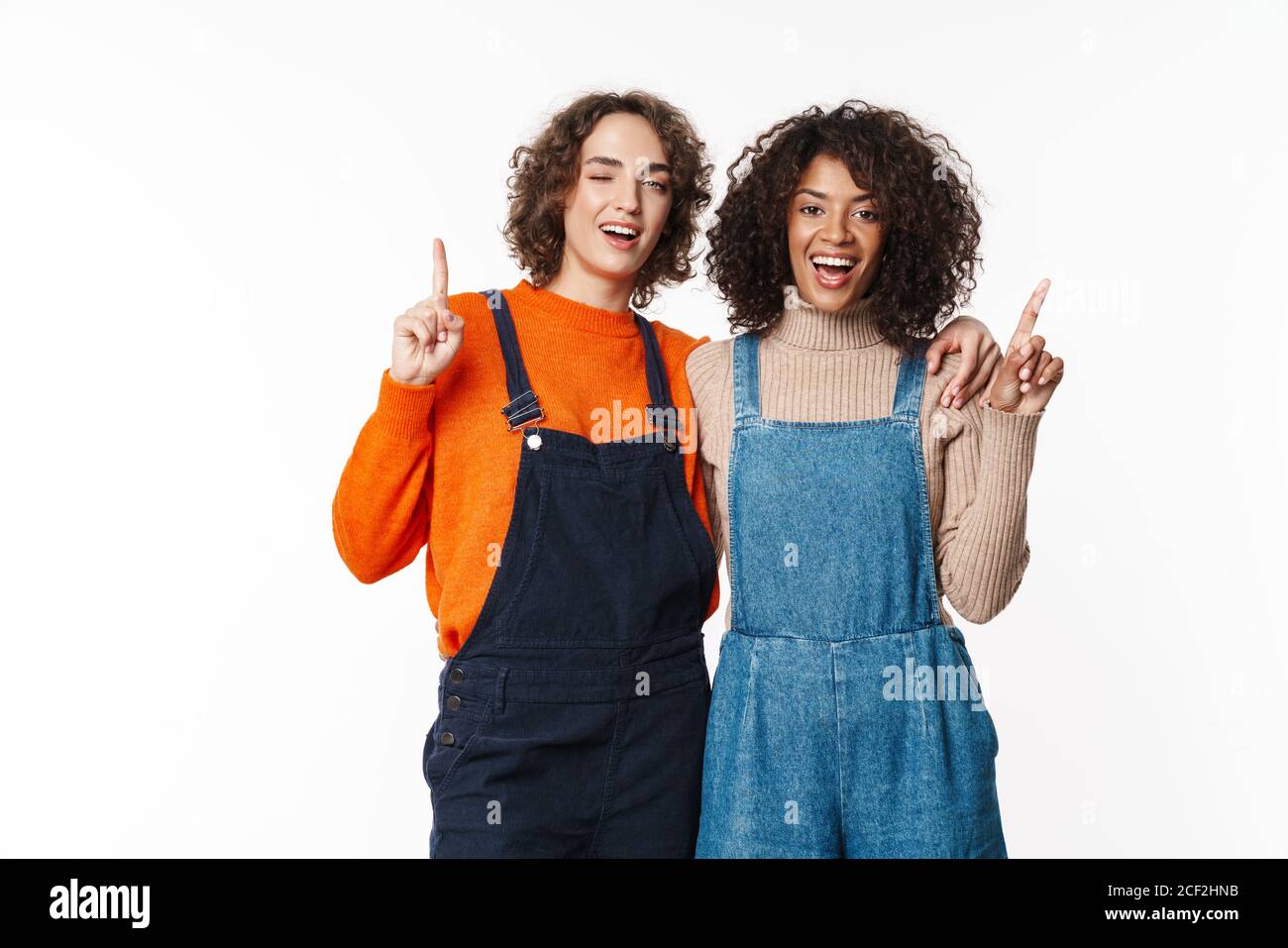 Image of cheery optimistic two multiracial girls friends in denim jumpsuits isolated over white wall background pointing to copyspace. Stock Photo