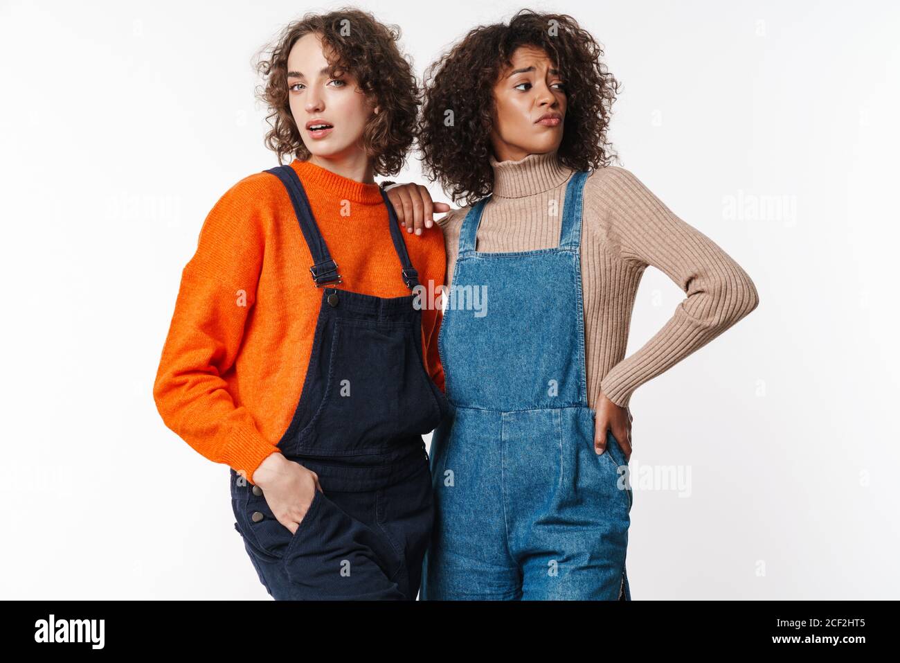 Portrait of confused multinational women in overalls posing and hugging ...