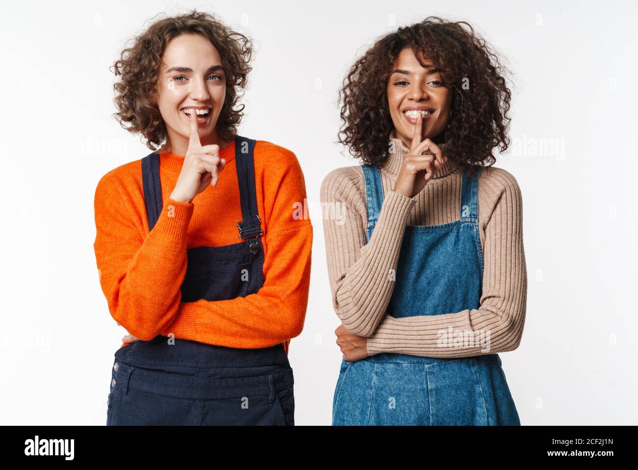Portrait of laughing multinational women in overalls gesturing silence ...