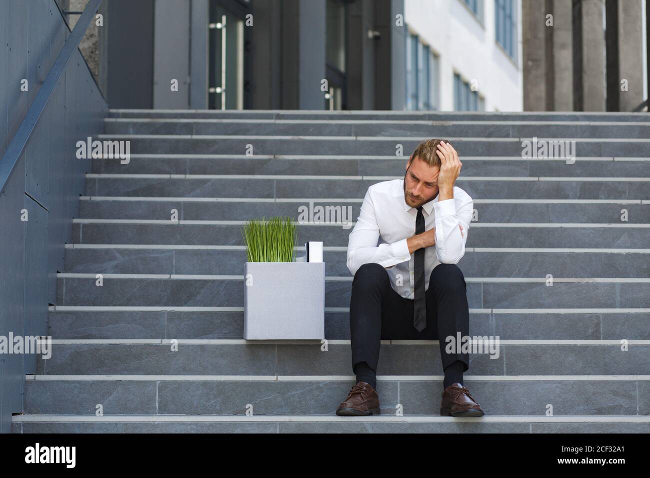 An unhappy young manager in a white shirt is fired from his job. A sad worker sits on the steps of a business center after layoffs. Crisis and Stock Photo