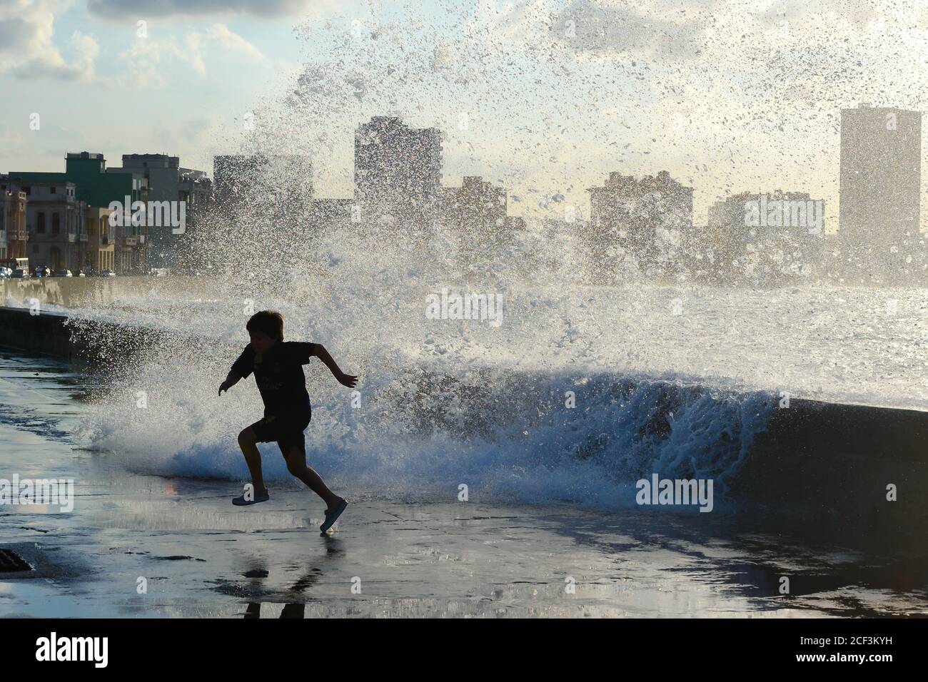 Cuban kid running from huge wave splash that hit the seawall in El Malecon, Havana, Cuba. Agitated sea hitting La Habana. Children having fun. Stock Photo