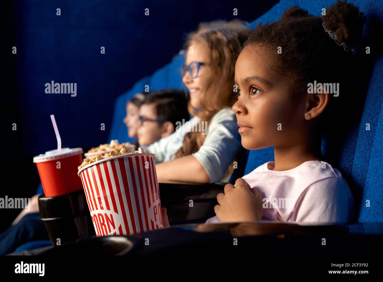 Beautiful concentrated african girl with funny hairstyle watching movie in cinema. Adorable little female kid sitting with friends, eating popcorn and smiling. Concept of entertainment, leisure. Stock Photo
