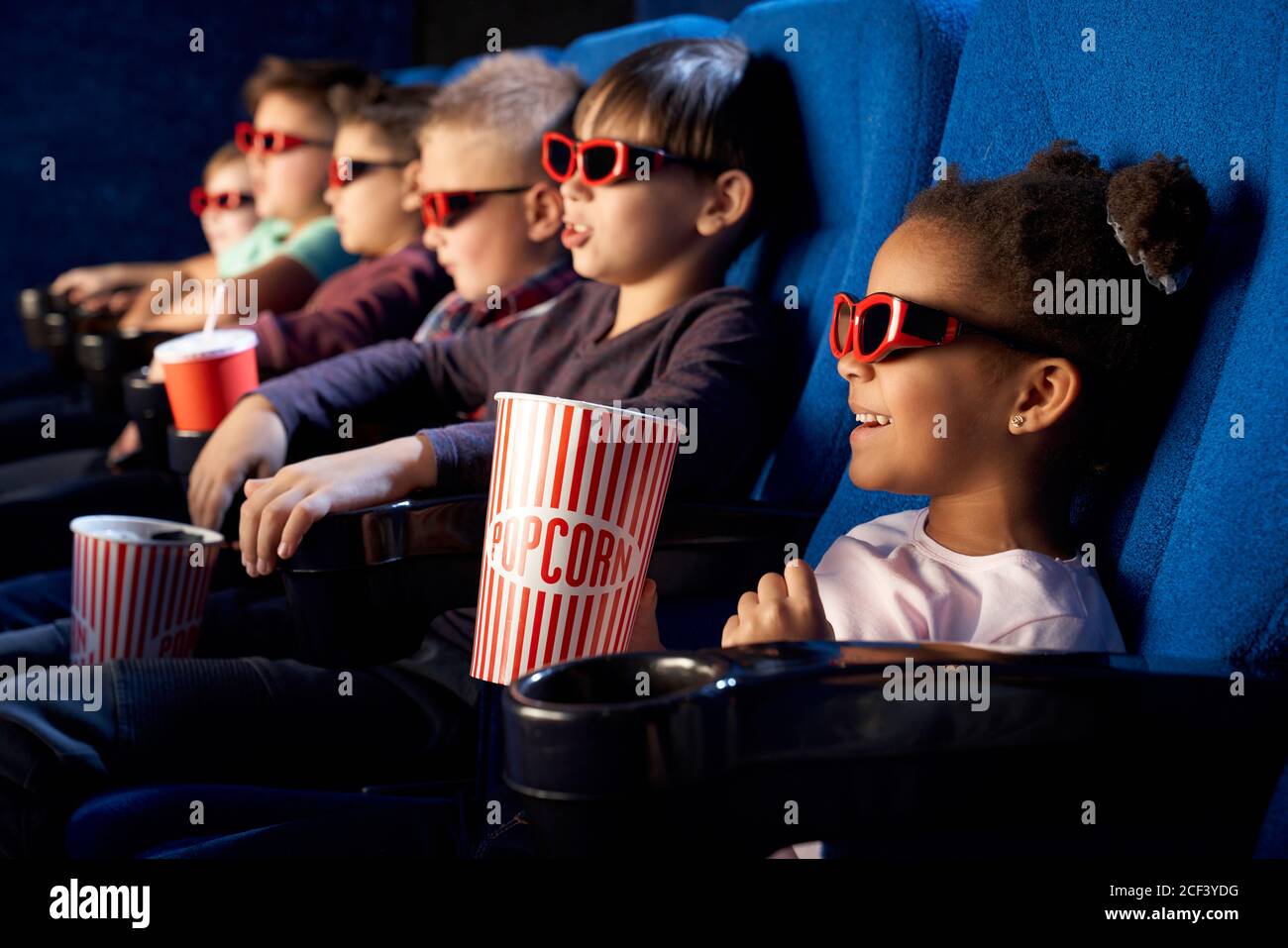Happy african female child wearing 3d glasses, holding popcorn and watching comical movie. Cute little girl spending free time with friends in cinema and laughing. Concept of leisure, entertainment. Stock Photo