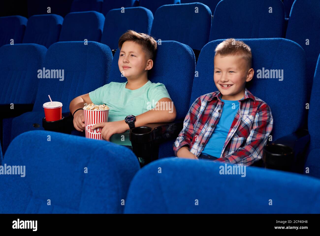 Side view of two smiling boys watching comical movie together in empty cinema. Male friends holding popcorn and sweet water. Children laughing and having rest on weekend. Concept of childhood. Stock Photo