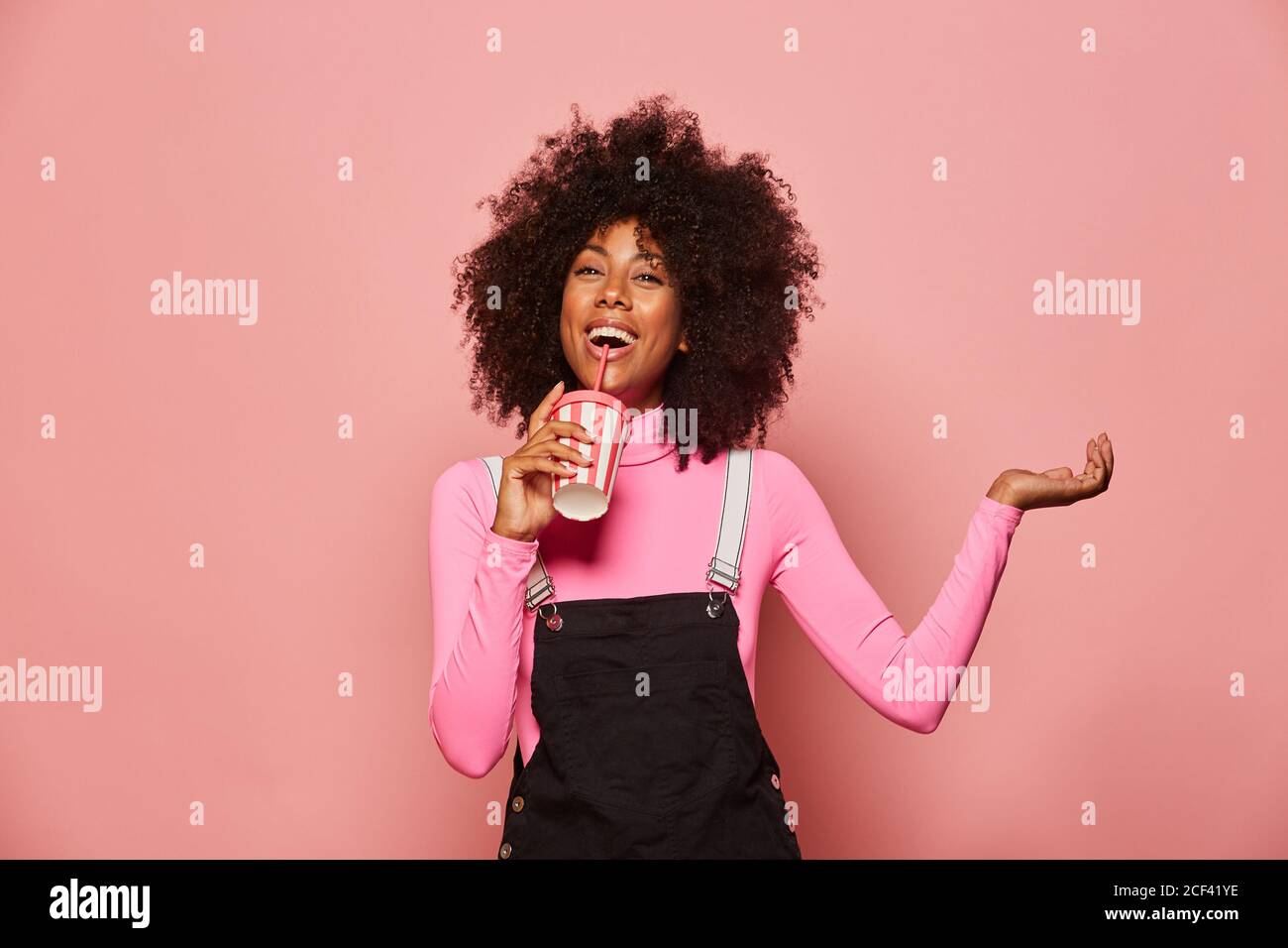 Joyful young African American female with curly hair wearing pink turtleneck and black overall holding white and pink striped disposable cup with straw and looking at camera Stock Photo