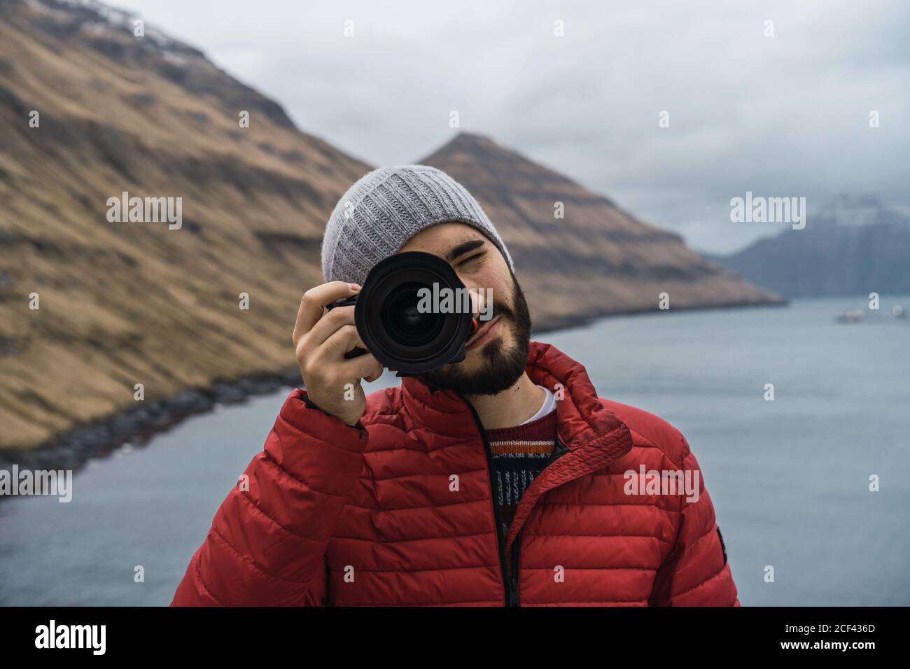 Bearded man in winter clothes pressing the shutter of a photo camera outdoors in Faroe Islands landscape Stock Photo