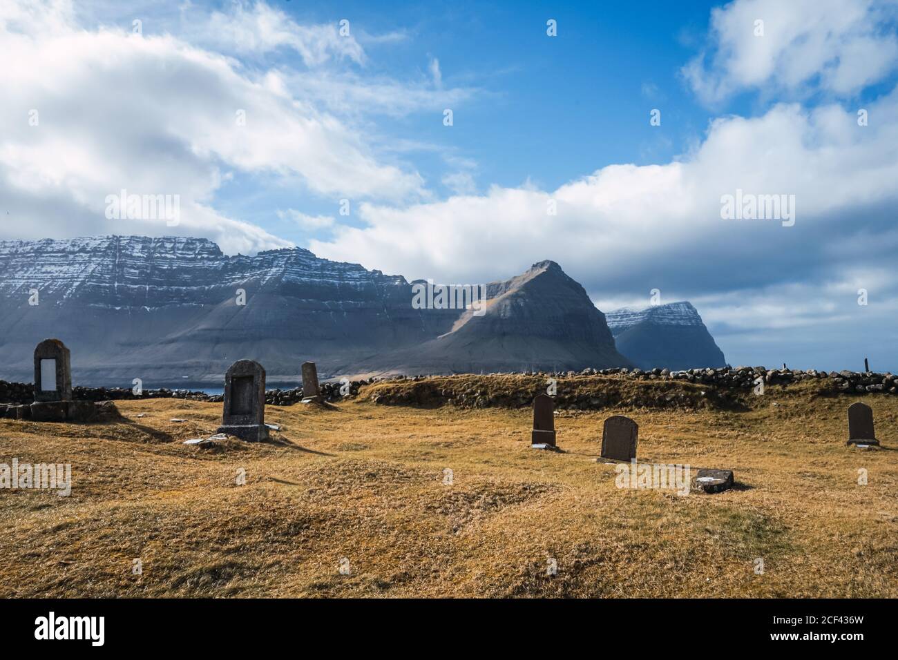 Ancient cemetery with old tombstones in field against backdrop of high mountains surrounding clouds on Faroe Islands Stock Photo