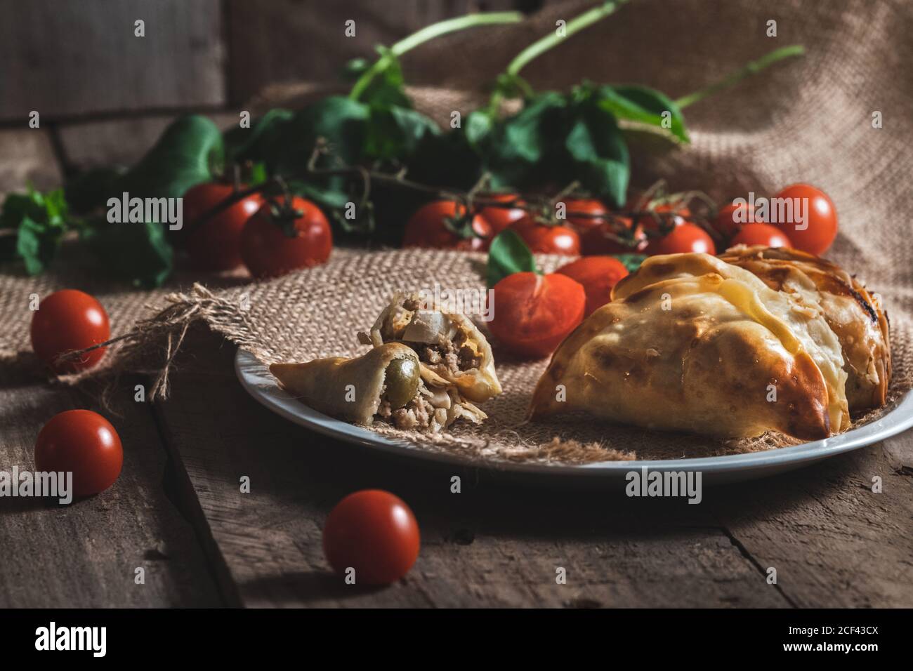 Appetizing freshly baked homemade turnover pies with meat and vegetable filling served on plate with ripe red tomatoes Stock Photo