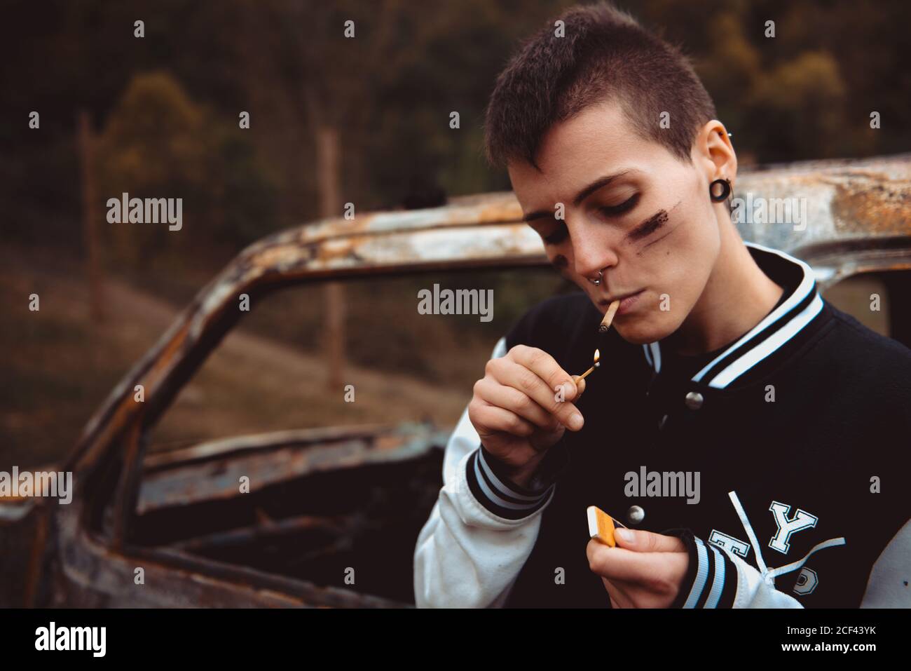 Young female with short hair and painted face lighting cigarette with lighter while standing near old rusty car in countryside and smoking Stock Photo