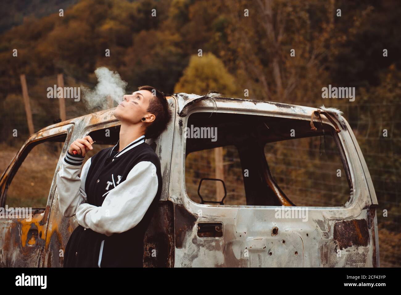Young Woman with short hair leaning on shabby burnt car and exhaling cigarette smoke while spending time in countryside Stock Photo