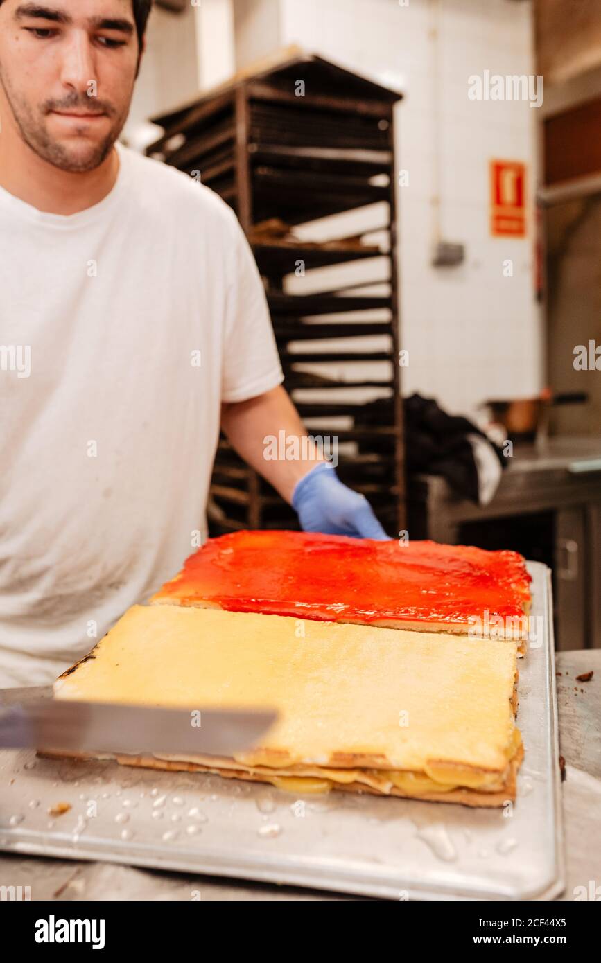 Crop man in white uniform holding tray with freshly glazed sweet cakes while working on blurred background of bakery kitchen Stock Photo