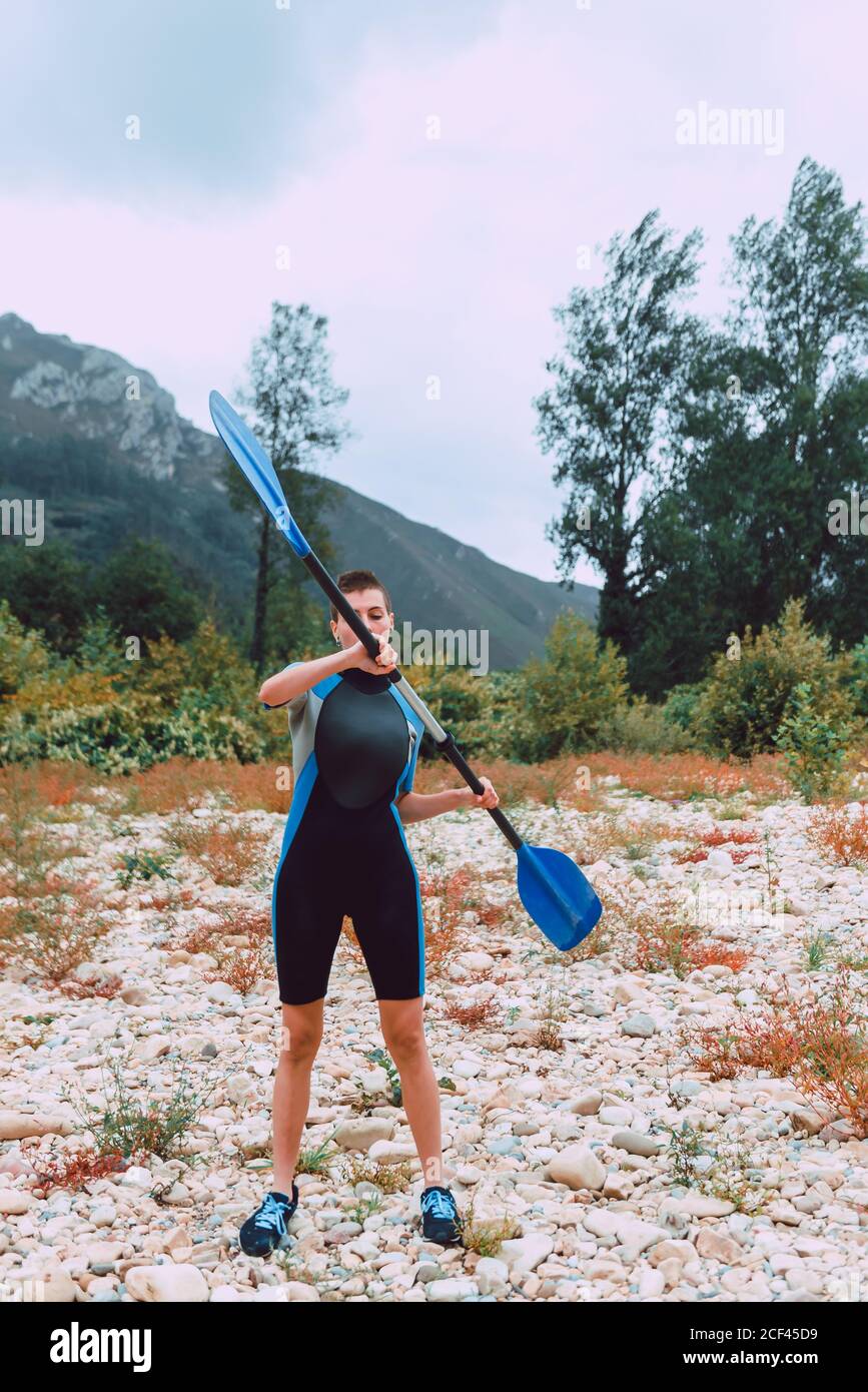 Sportive Woman in wetsuit training with paddle while standing on bank of Sella river covered with limestones and looking at camera in Spain Stock Photo