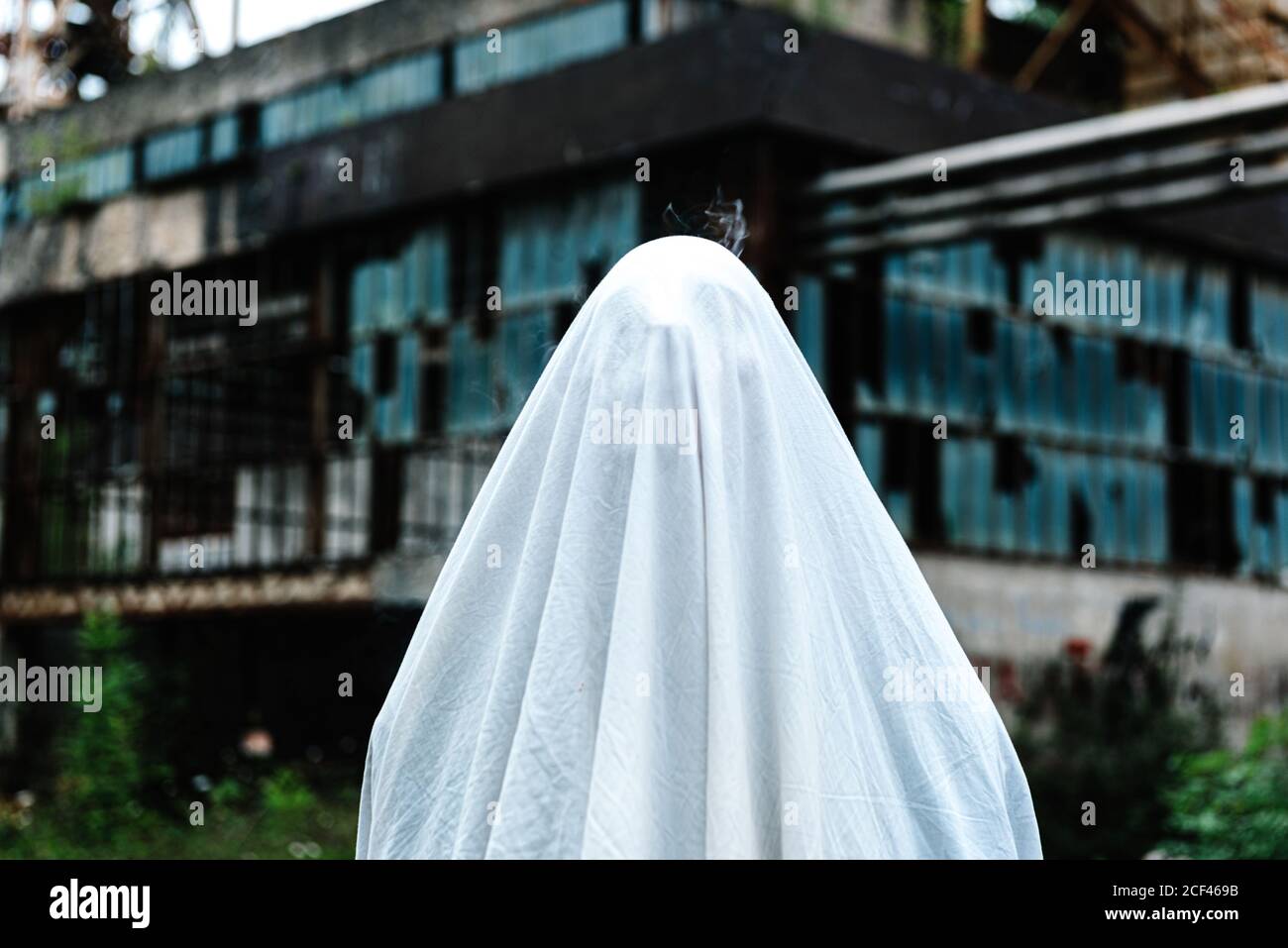 unrecognizable person covered in white bed sheet masked as a ghost near old abandoned mine building with rusty metal construction and shabby walls Stock Photo