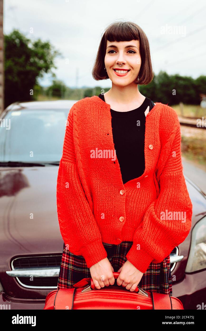 Charming attractive Woman with stylish hair dress in red sweater with red suitcase waiting for trip in car behind on road Stock Photo