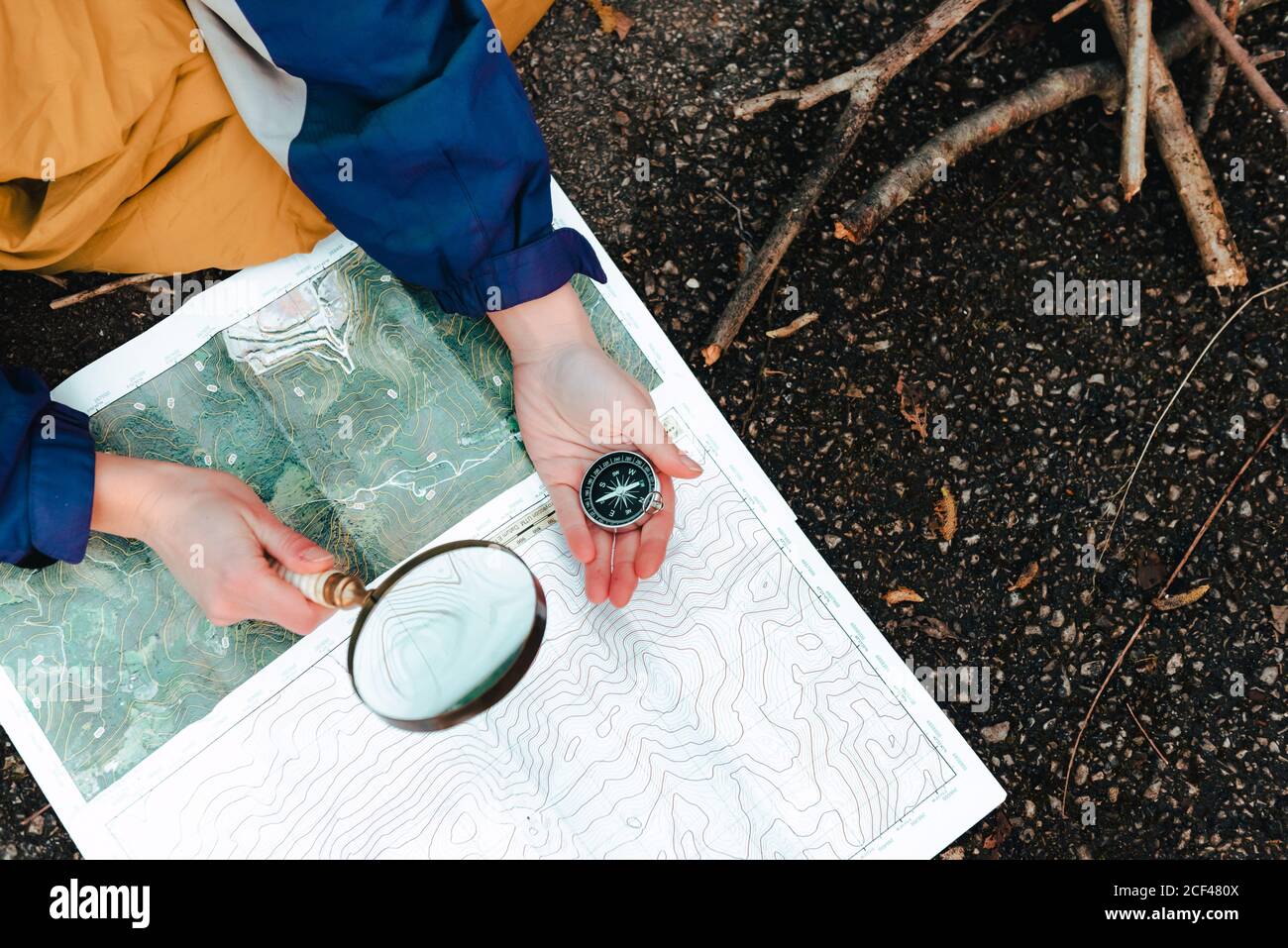 From above crop traveler checking route on topographic map with compass and magnifying glass while sitting on ground in autumn day Stock Photo