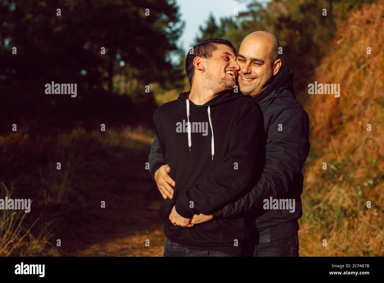 Cheerful homosexual couple embracing on walkway in forest in sunny day on blurred background Stock Photo