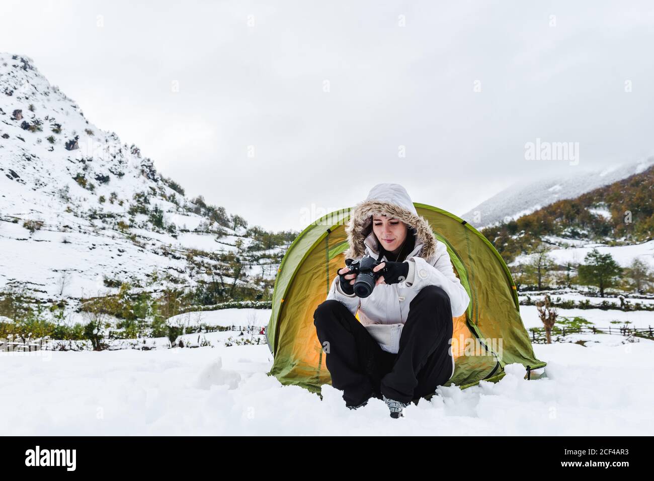 Content female in white winter jacket and black pants sitting with camera near light green tourist tent on snow in mountains Stock Photo