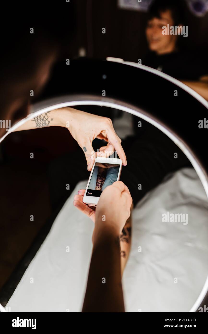 unrecognizable man tattooer using smartphone to take picture of tattoo on leg of crop customer for portfolio in contemporary studio Stock Photo